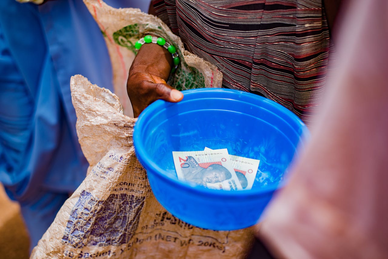 about-us A close-up of a hand holding a blue bowl containing currency notes outdoors.